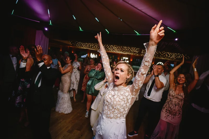 Party bride in jumpsuit puts her hands in the air on the dance floor at elmore court