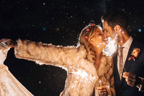 Boho bride in white lace wedding dress, white fur coat and flower crown holds her dress out to the side in one hand and a sparkler in the other as she leans in to kiss her modern groom in navy suit and patterned tie who is also holding sparkler at romantic modern boho wedding at night at wild wedding venue elmore court