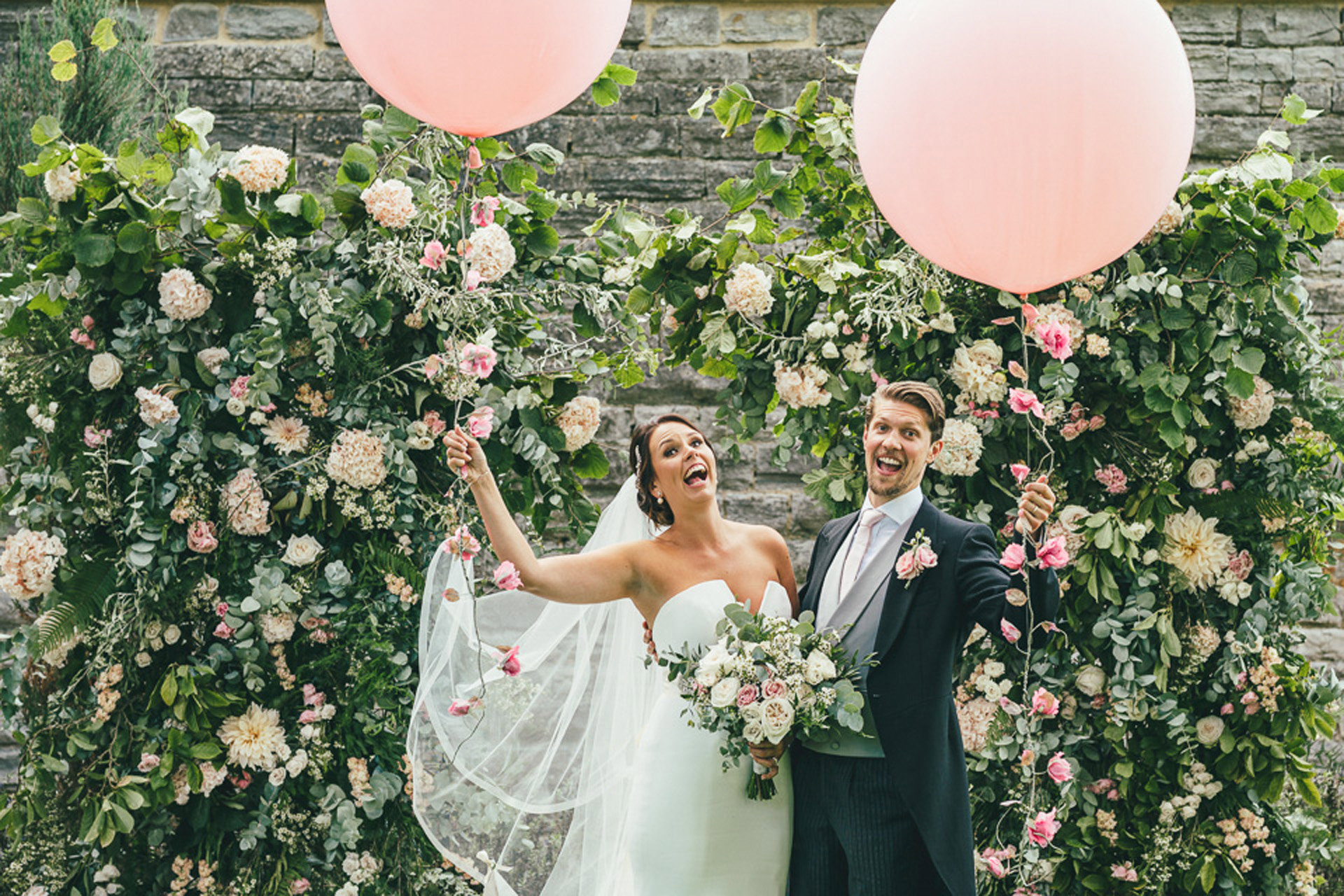 Fun newlywed couple pose with giant pink balloons in front of beautiful romantic wedding arch of pink and white roses
