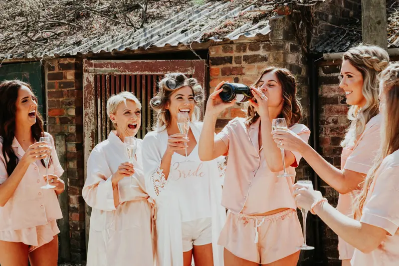 Bridal party in pink and white robes and pyjamas laugh as the bride drinks prosecco out of the bottle