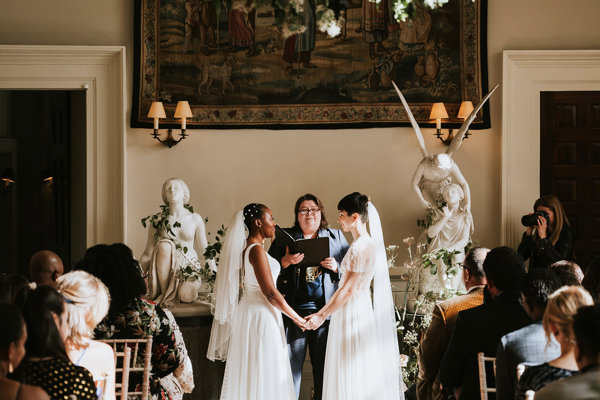 Two brides stand facing each other holding hands in their beautifully atmospheric wedding ceremony in the hall of stately home in the cotswolds