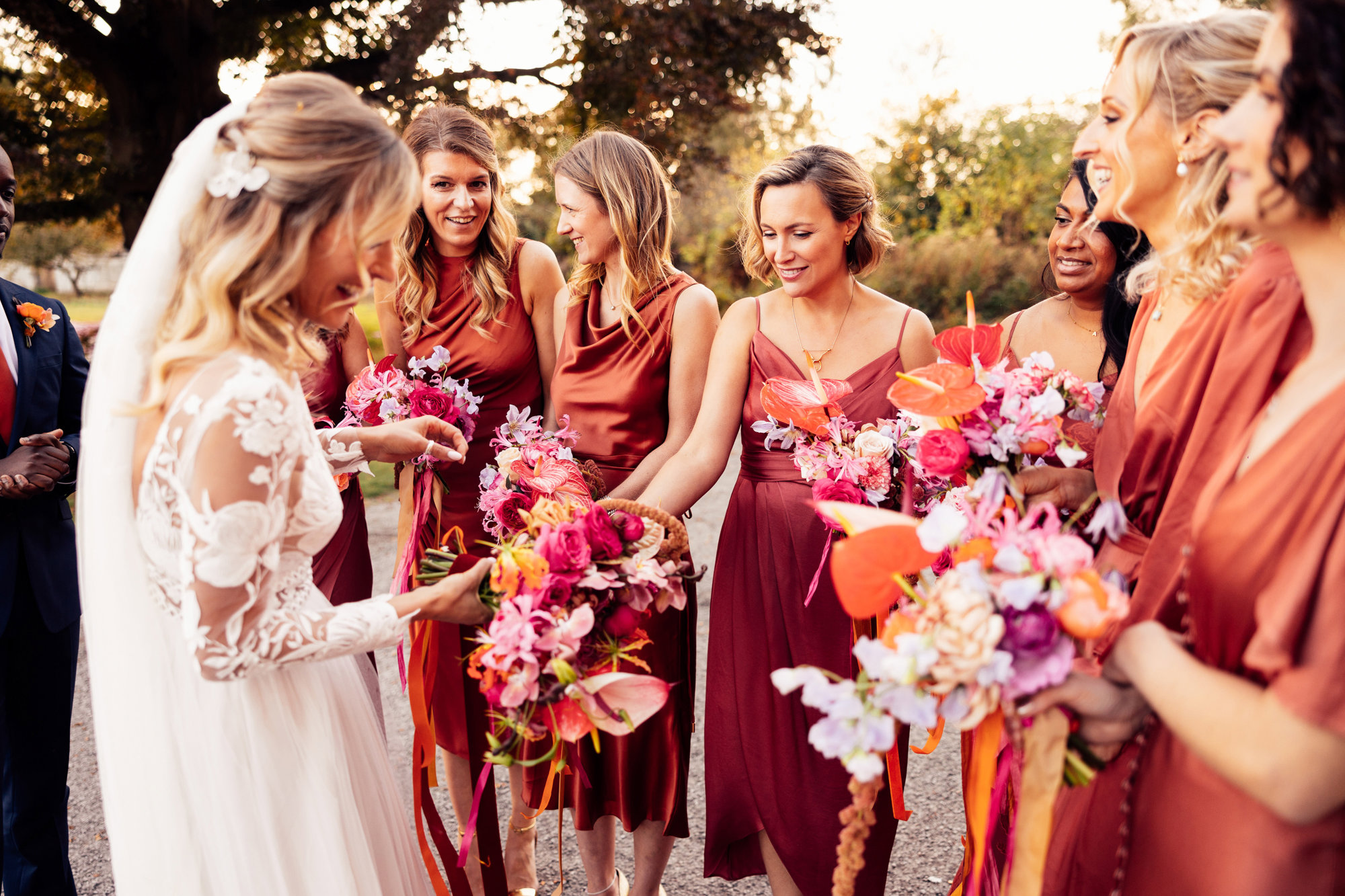 Colourful bridesmaids in orange dresses stand with boho bride with bright wedding flowers with ribbons in the cotswolds countryside