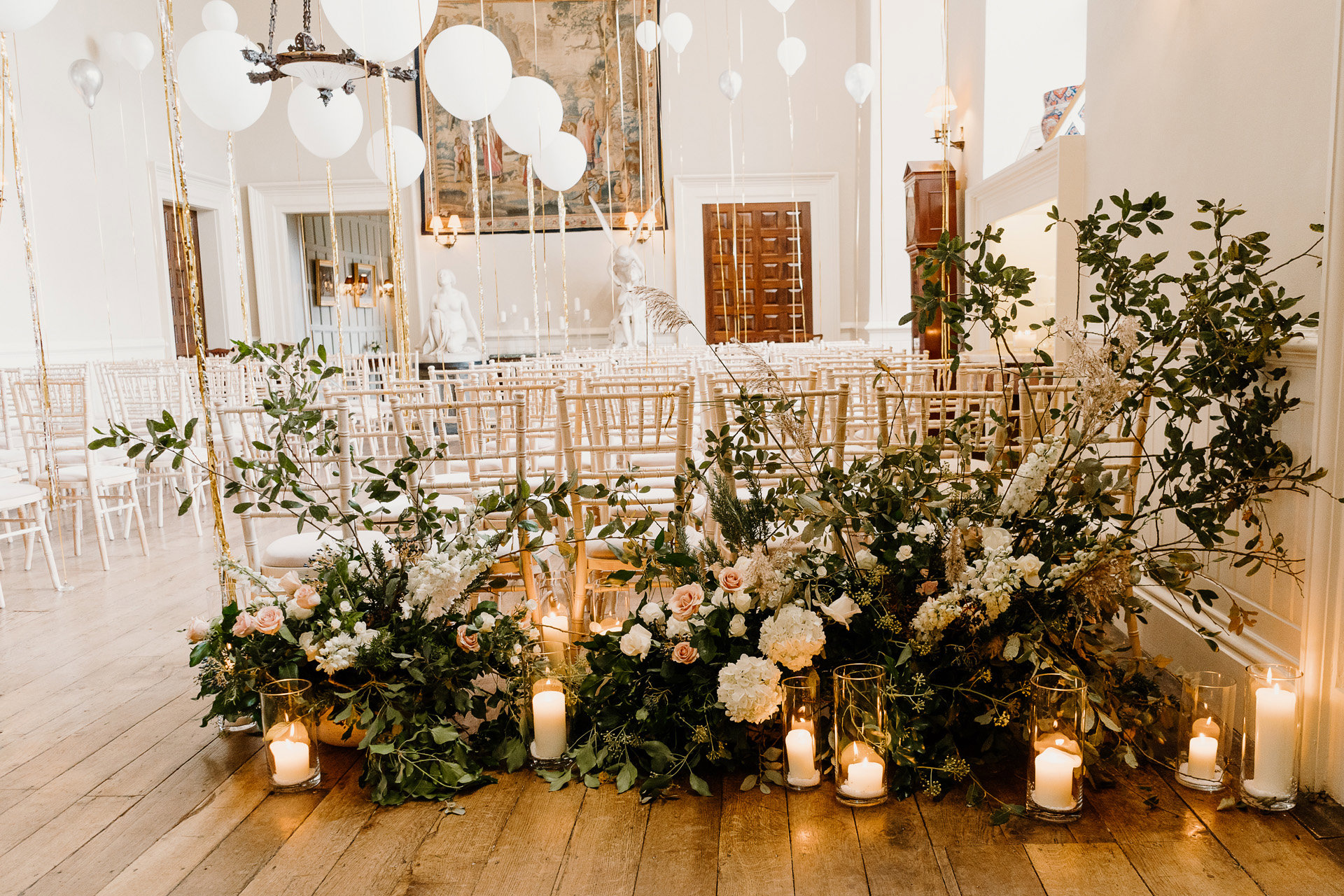 Romantic wedding aisle styling with candles, white balloons and white flowers and greenery in a stately home hall