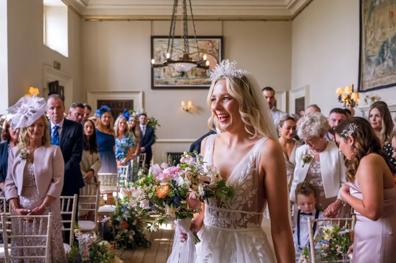 Beautiful blonde boho bride in pearl crown and lace and tulle wedding dress holding bright bouquet at the end of aisle with guests standing behind in historic hall of elmore court