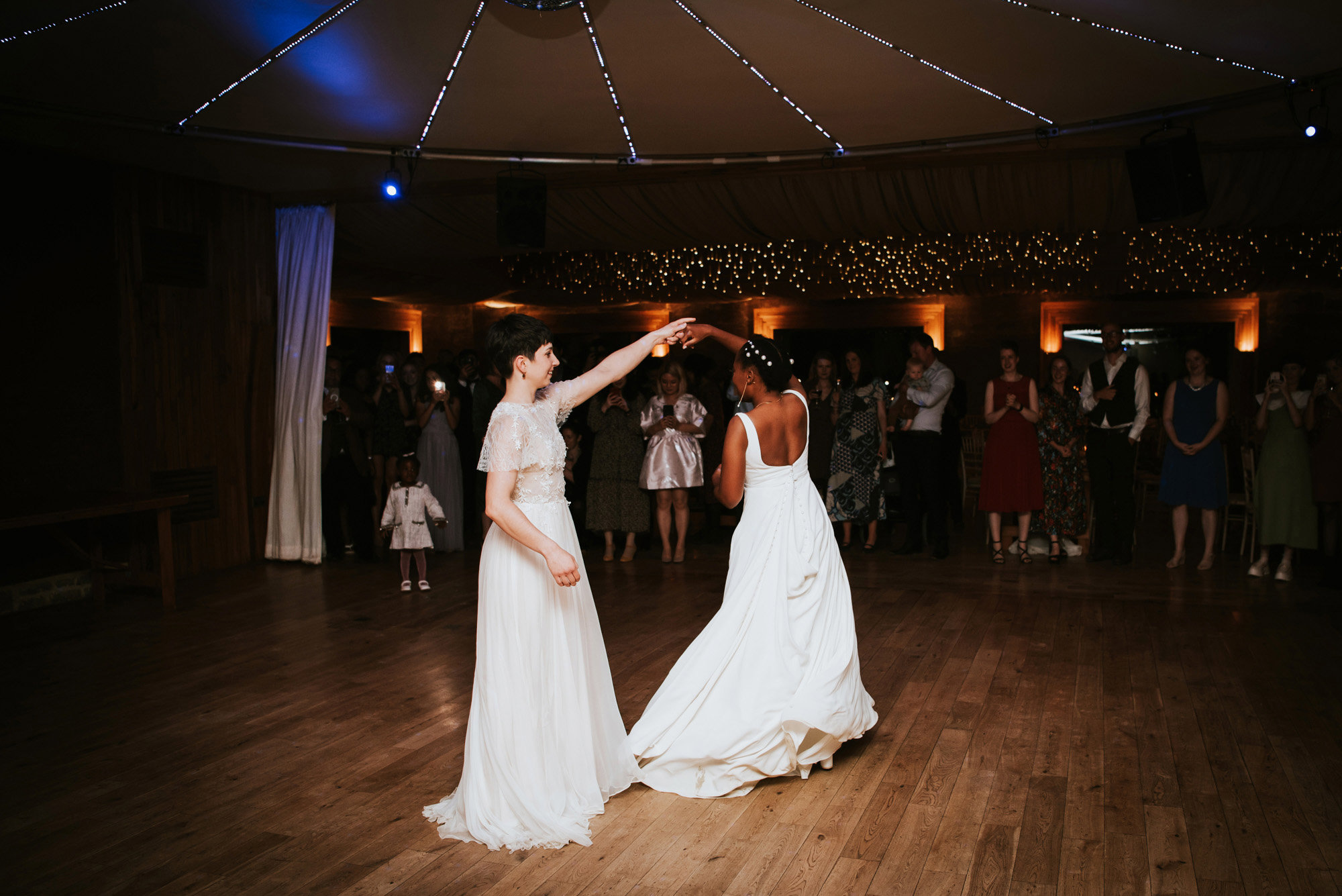 Two brides dance together on the dancefloor at eco party venue in white wedding dresses at their romantic modern multicultural Lesbian wedding
