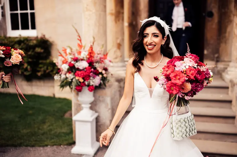 Beautiful modern bride in pearl accessories; headpiece and pearl necklace holding bright pink and red bouquet and pearl handbag outside her glamorous wedding venue