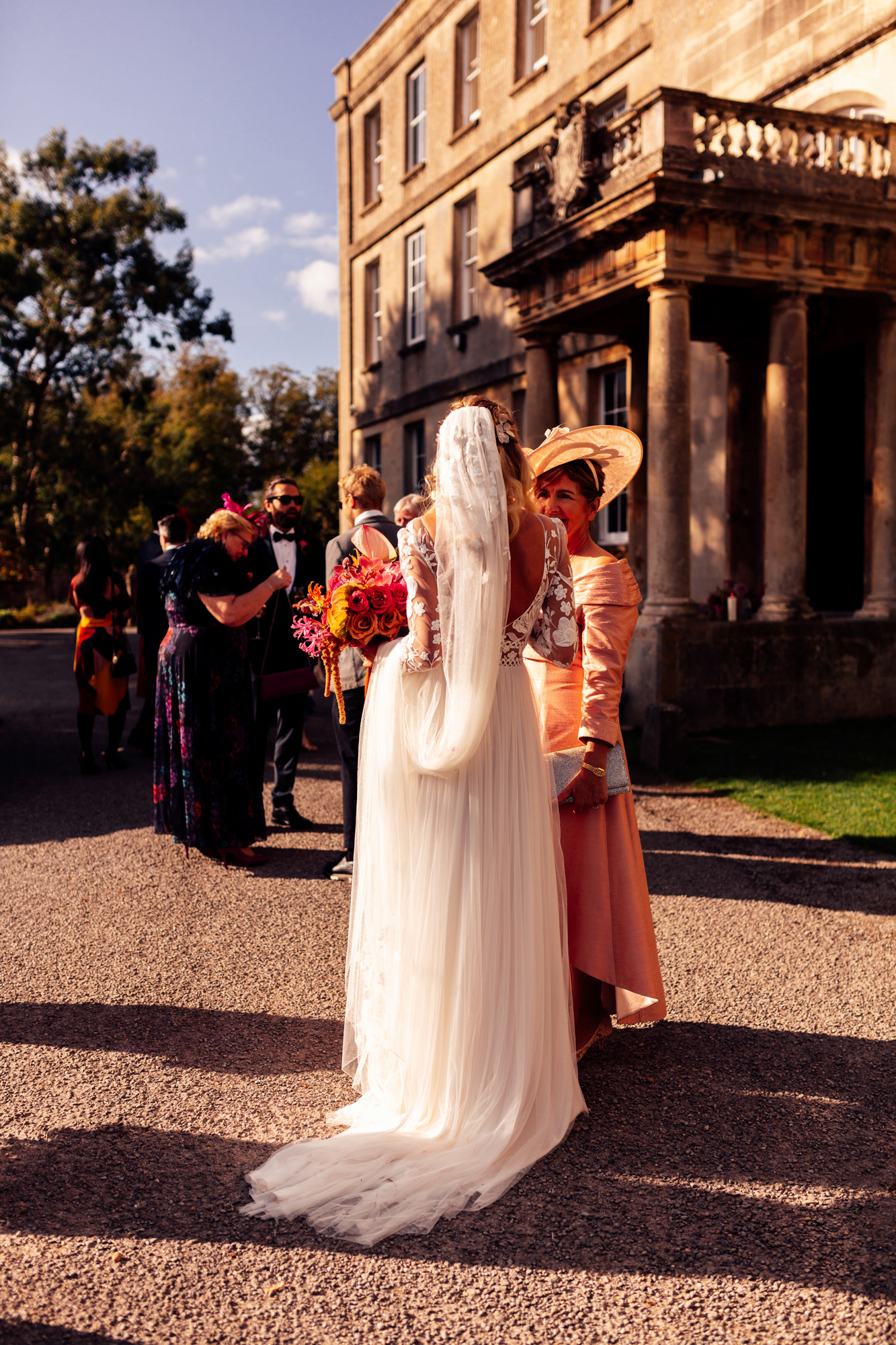 Cool modern boho bride in lace dress and long veil holding bright colourful wedding bouquet in the sunshine outside stately home party wedding venue in the cotswolds