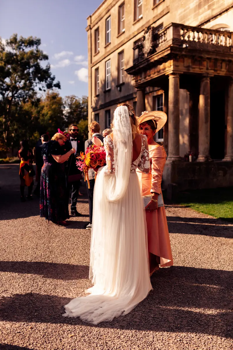 Cool modern boho bride in lace dress and long veil holding bright colourful wedding bouquet in the sunshine outside stately home party wedding venue in the cotswolds