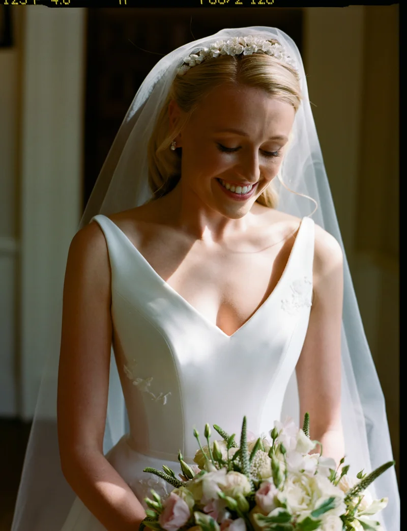 Demure and elegant bride in chic simple satin wedding gown, pearl tiara and veil, pearl necklace and pearl earrings smiles down at her bouquet of white, pale pink and green flowers