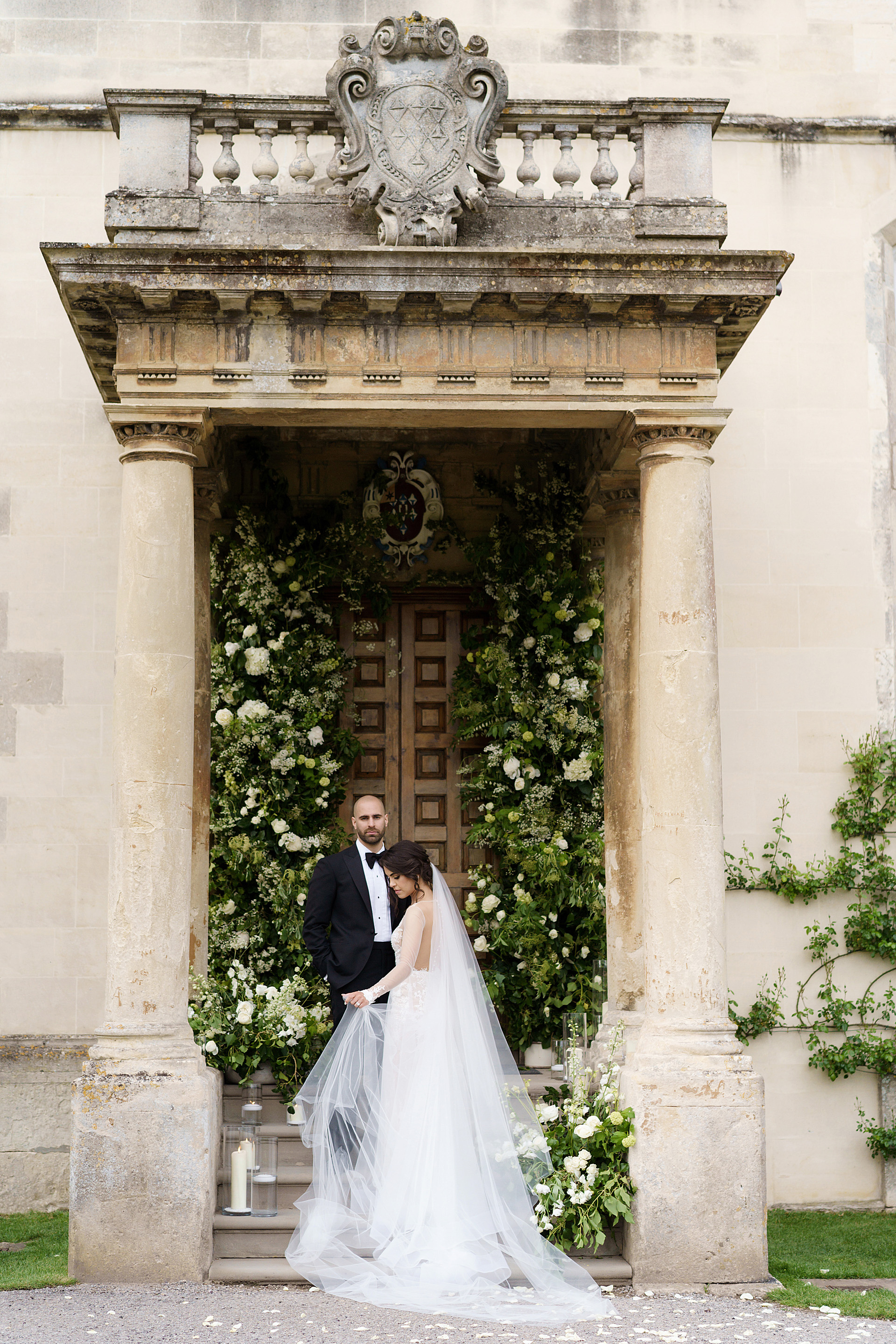 bride and groom stand under porch with columns against backdrop of huge statement greenery and white floral arrangement for a micro wedding