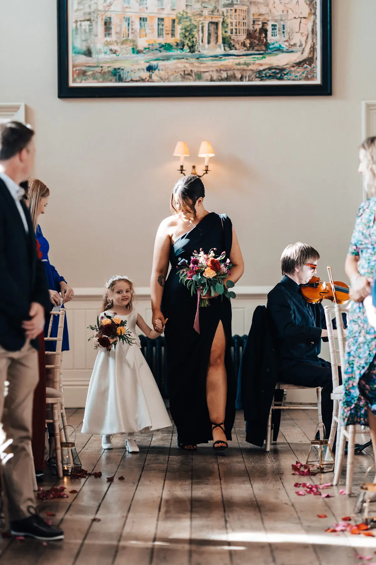 bridemaid wearing black dress walks down aisle holding flower girls hand in an Autumn wedding ceremony at a country house in gloucestershire