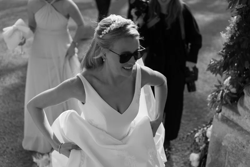 Cool modern bride in Ray Ban sunglasses, pearl bridal tiara, pearl necklace and pearl earrings lifts up the skirt of her princess wedding dress to walk up the steps of stately home wedding venue