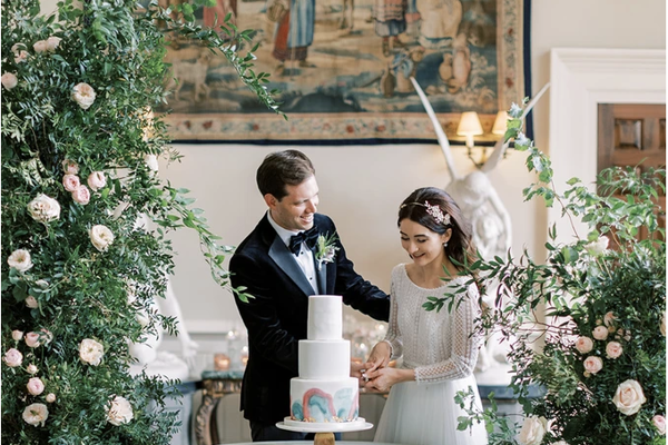 Luxe bride and groom cut rainbow cake surrounded by greenery flower arches in stately home wedding venue in the UK