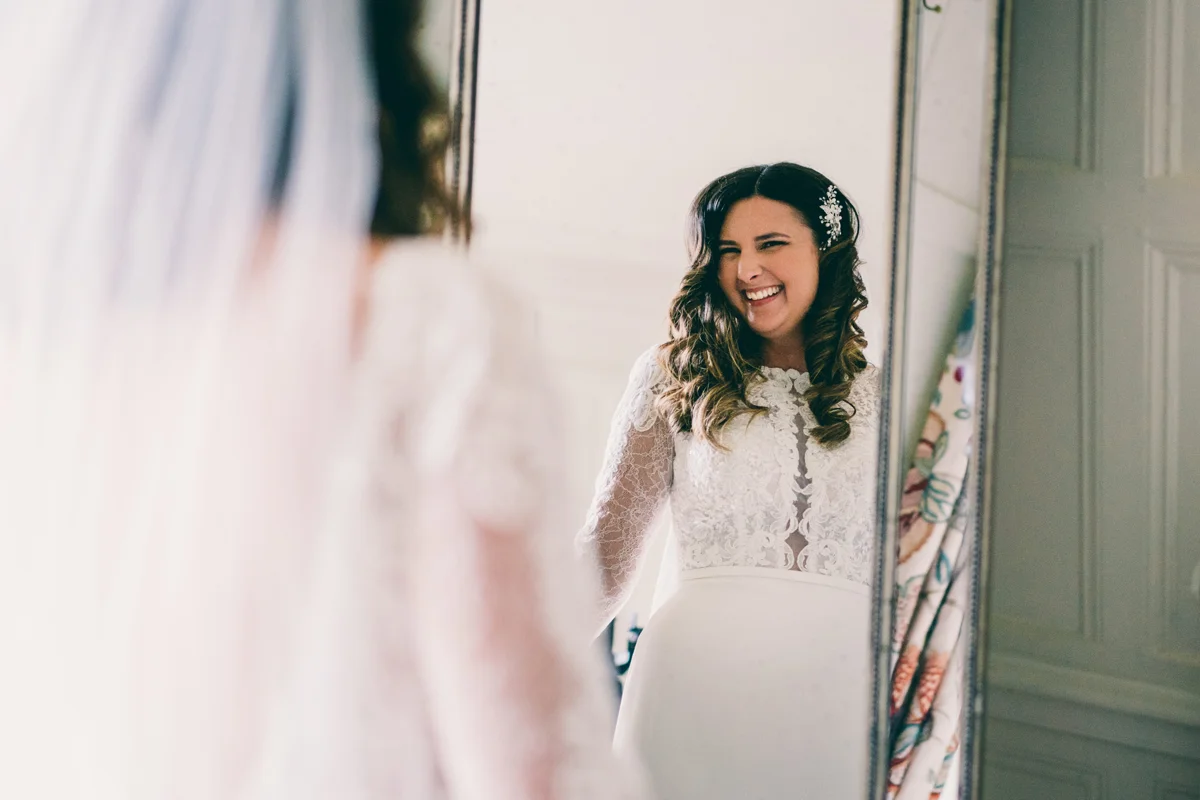 Bride in lace dress smiles in mirror at brunette curly hair with pearl hair clip