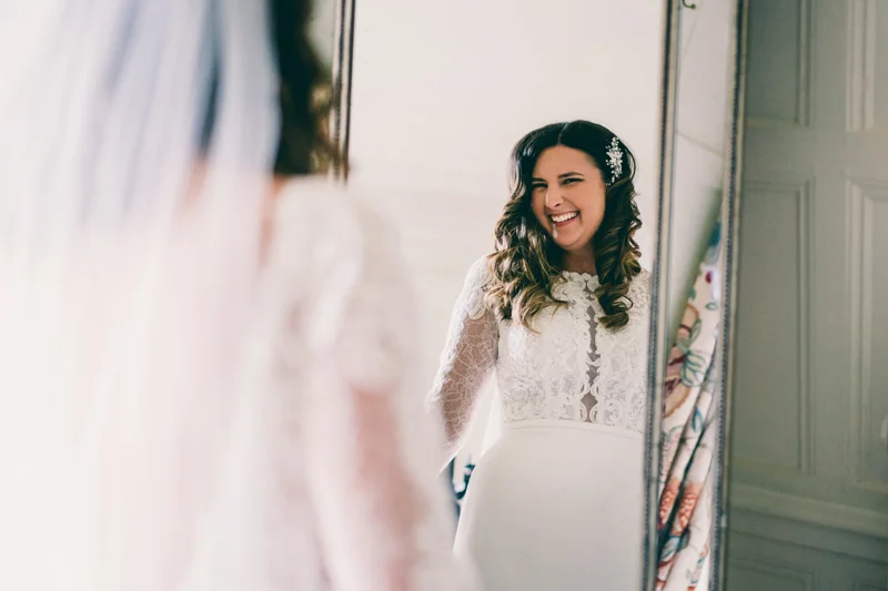 Bride in lace dress smiles in mirror at brunette curly hair with pearl hair clip