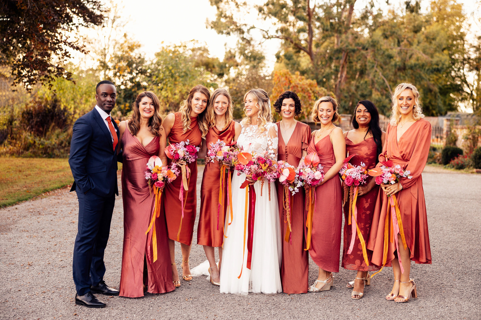 brides man and bridesmaids in burnt orange copper dresses stand with boho bride with bright bouquets outside stately home wedding venue in the cotswolds countryside