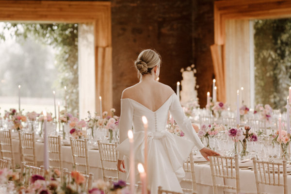 A modern bride in stunning scoop back wedding dress with huge bow surveys her gorgeous romantic soft pink wedding reception with pretty flowers and candles laid out on glowing tables in the twinkling light of The Gillyflower at a modern wedding at Cotswolds venue Elmore Court