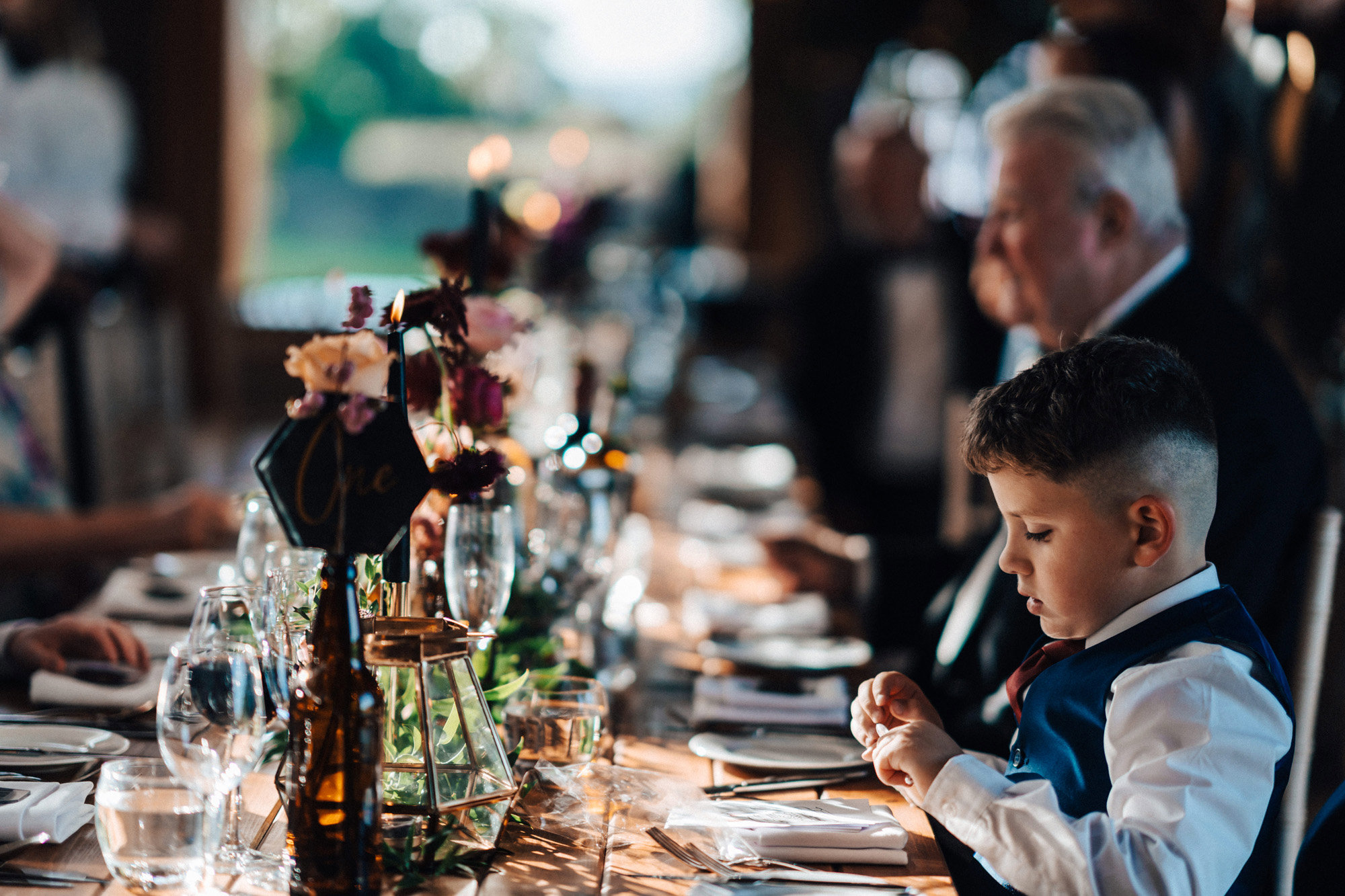 Wedding guests sitting at long banquet tables decorated with black flowers and mismatched glasswear for a dark and moody autumn wedding in the cotswolds
