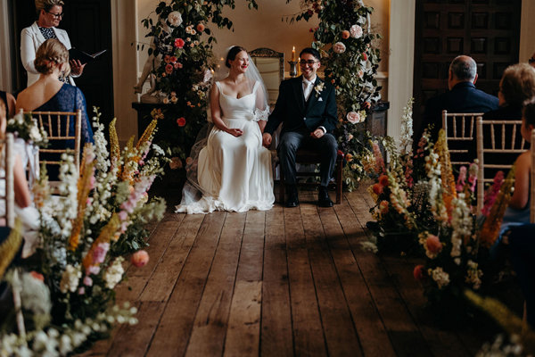 Beautiful newly wed couple sitting in front of a flower arch in the Great Hall at Elmore Court