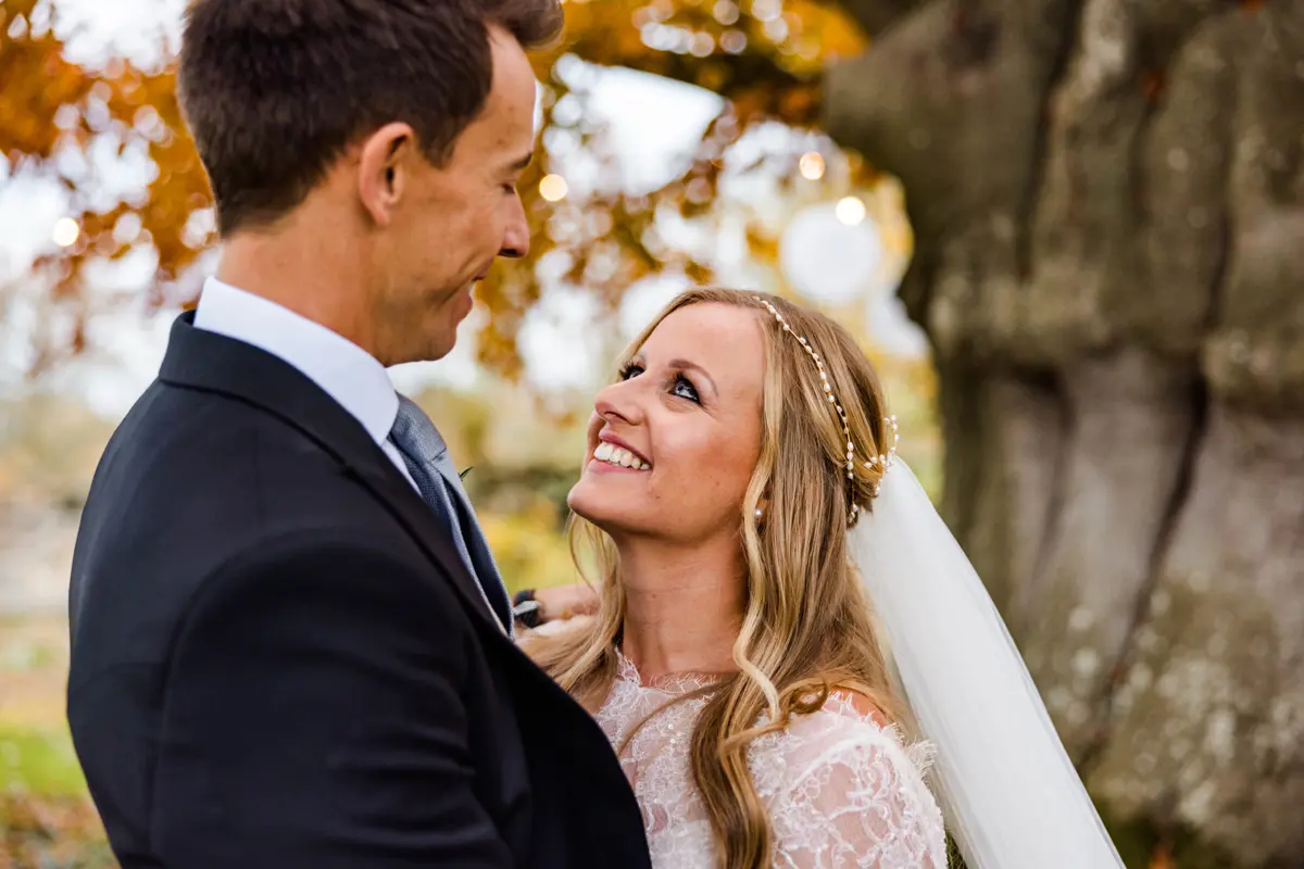 Bride in pearl headpiece and veil and groom stand underneath copper beech with golden leaves look lovingly at eachother