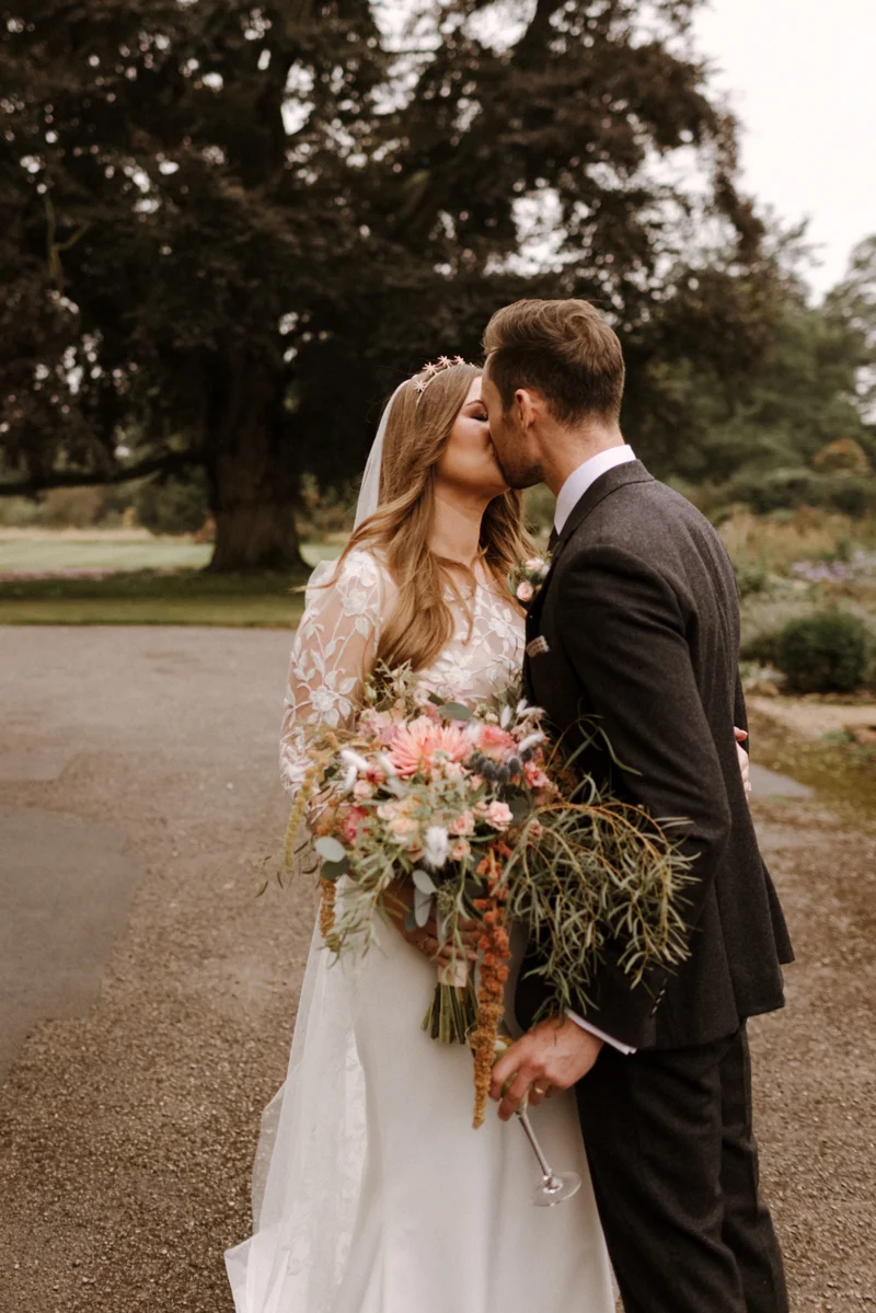 Beautiful boho bride kisses groom holding whimsical bouquet of trailing pink flowers at eco venue elmore court