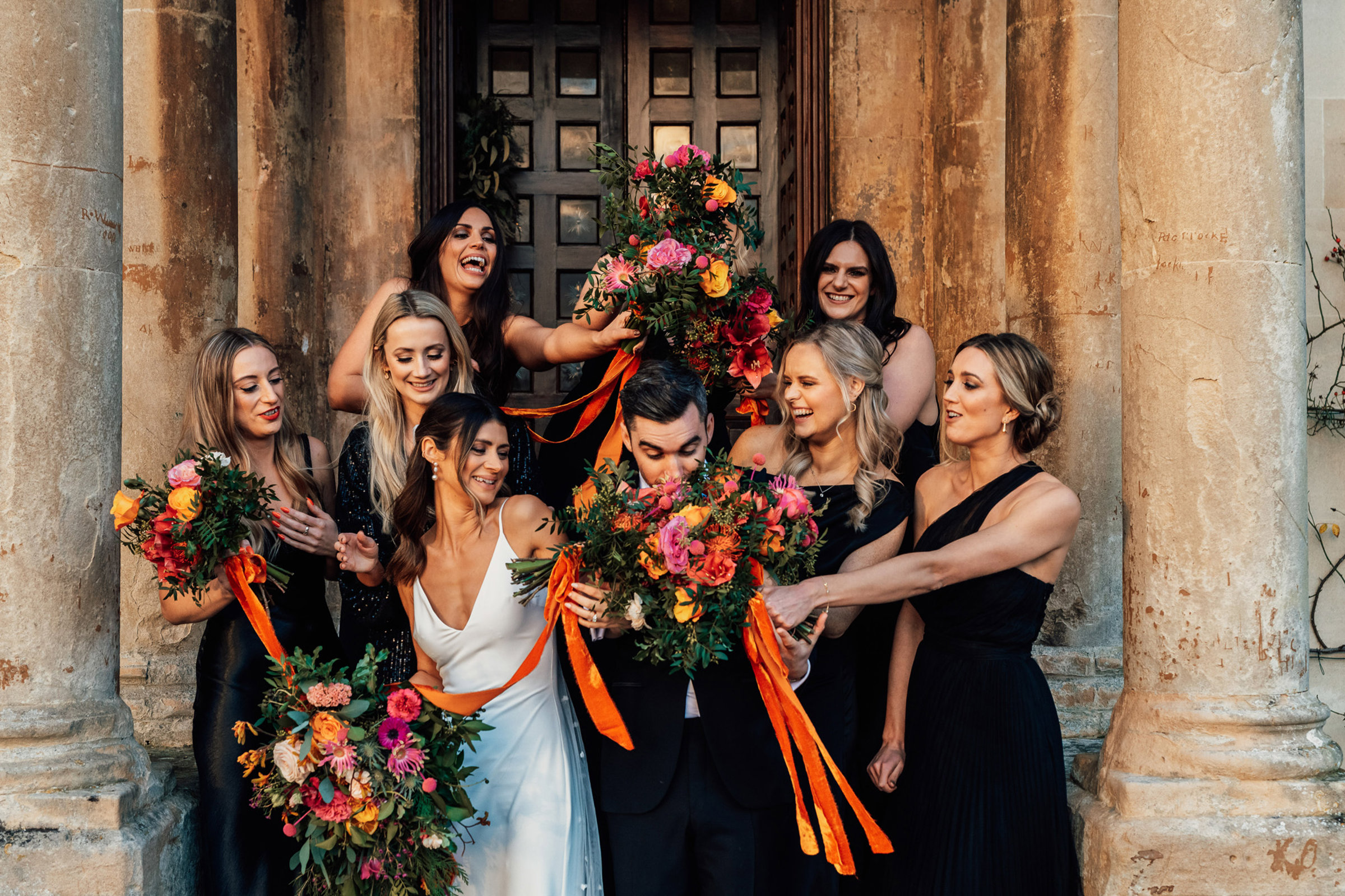 Cool couple pose on steps of stately home at their Black tie winter party wedding with bridesmaids in black and bright bouquets with long ribbons