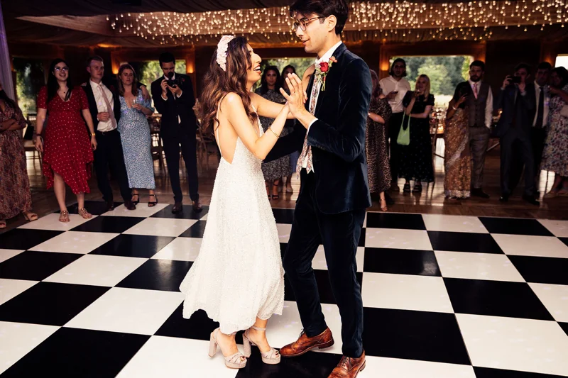 Modern bride wearing pearl reception dress and groom in blue velvet suit dance on chequered dance floor with twinkling lights above
