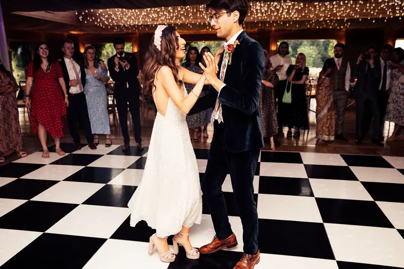 Modern bride wearing pearl reception dress and groom in blue velvet suit dance on chequered dance floor with twinkling lights above