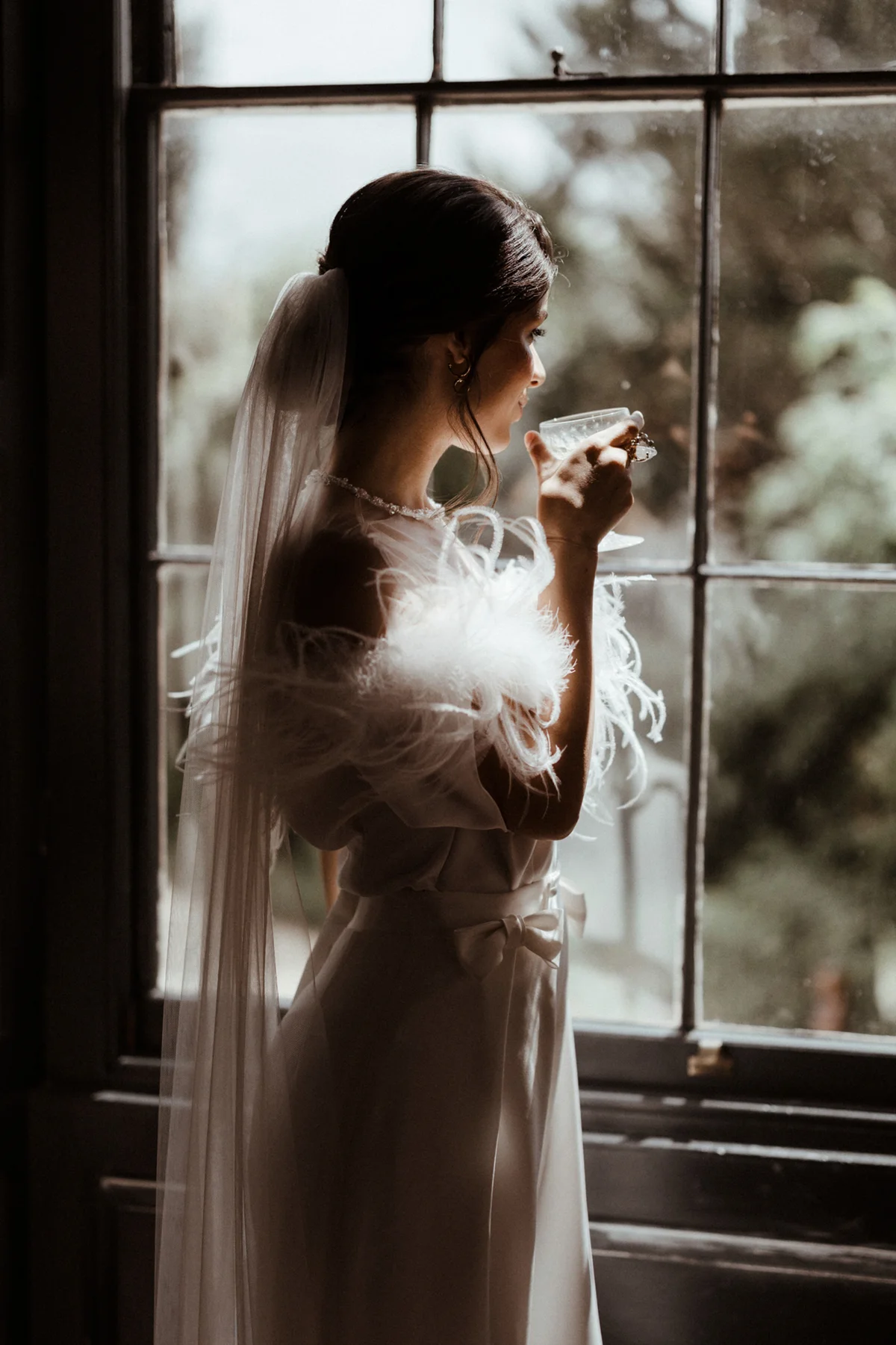 Glamorous parisian bride in pearl necklace, feathers and long veil and chignon bun holds crystal champagne glass looking out stately home wedding venue window