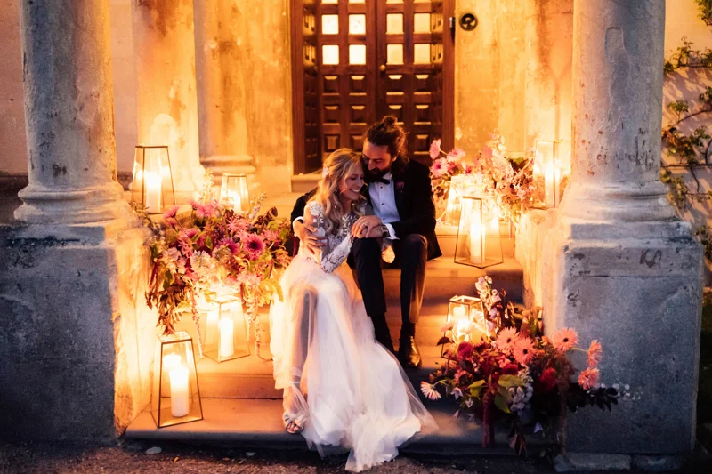 Bride and groom embrace on the steps of a stately home in Gloucestershire, surrounded by glowing candle light and stunning flower arrangements