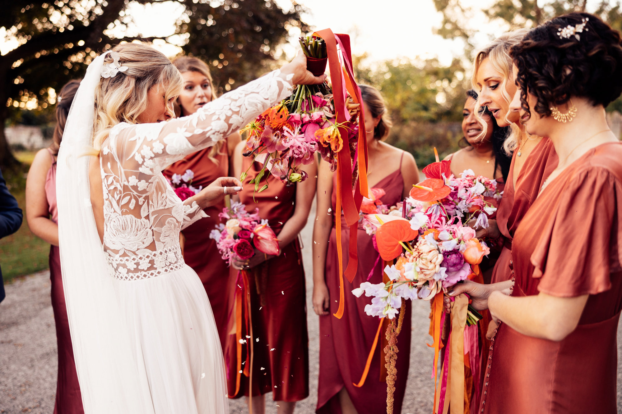 Colourful bridesmaids in orange dresses stand with boho bride with bright bouquets with ribbons in the cotswolds countryside
