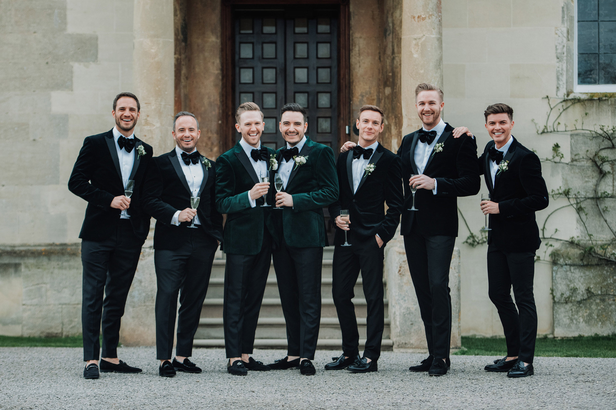 Gay wedding party 2 grooms and their 5 groomsmen standing outside their stately home wedding venue elmore court wearing matching velvet tuxedos