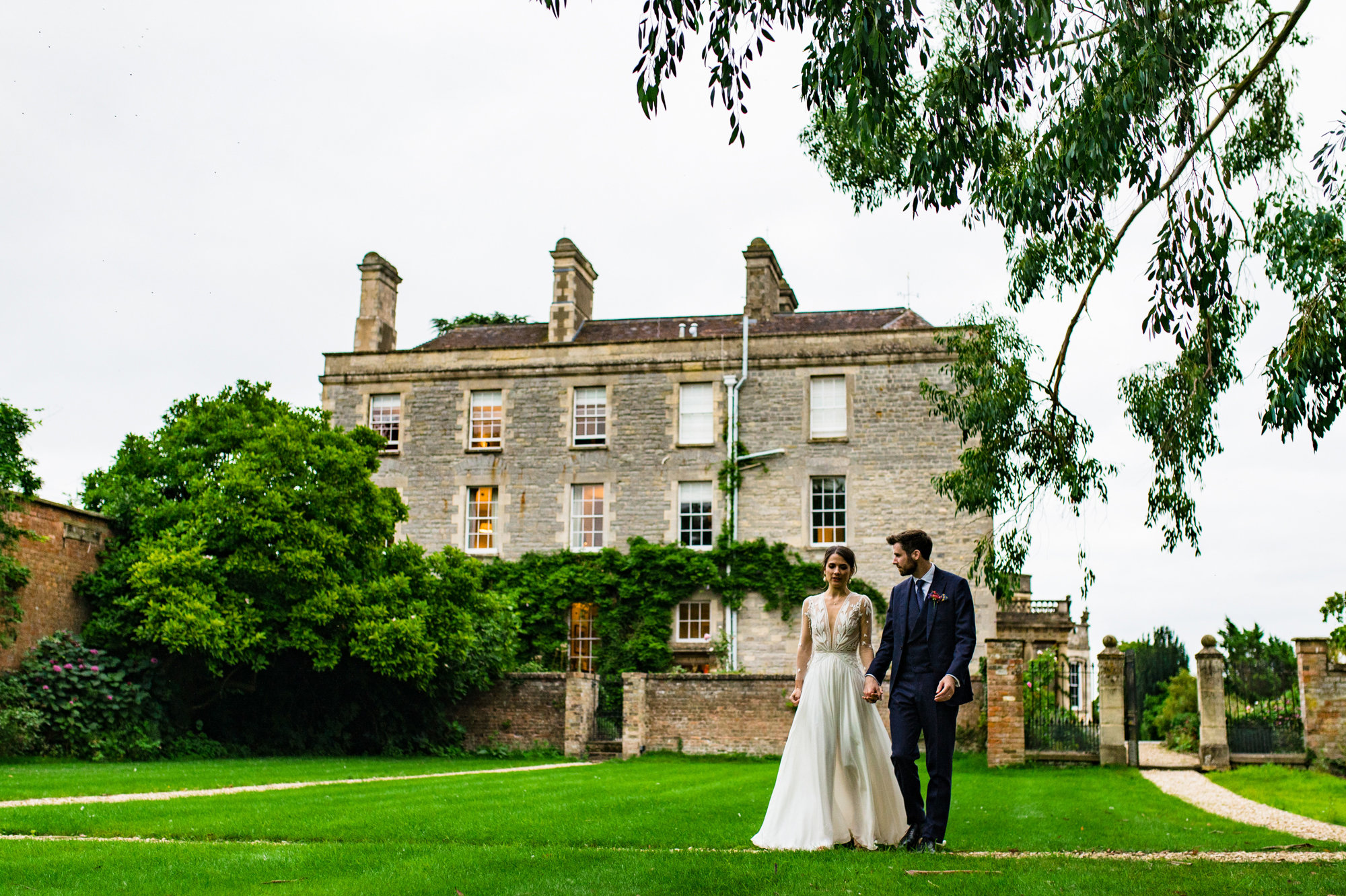 elegant bride and groom walk through the walled garden hand in hand with stately home elmore court in the background