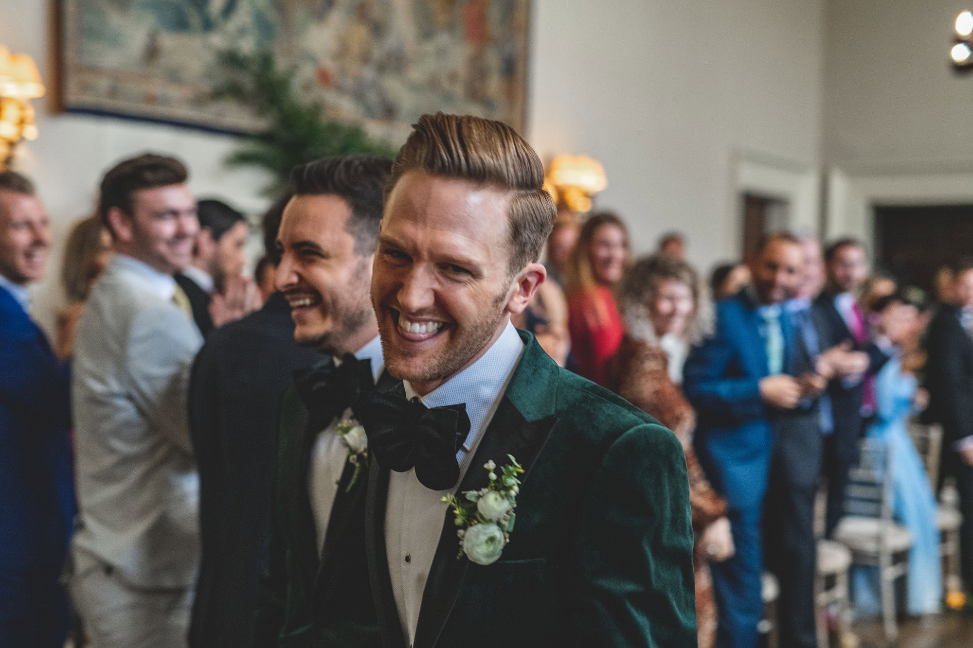 Handsome groom laughs at the camera walking down the aisle with his new husband wearing matching tuxedos at same sex ceremony