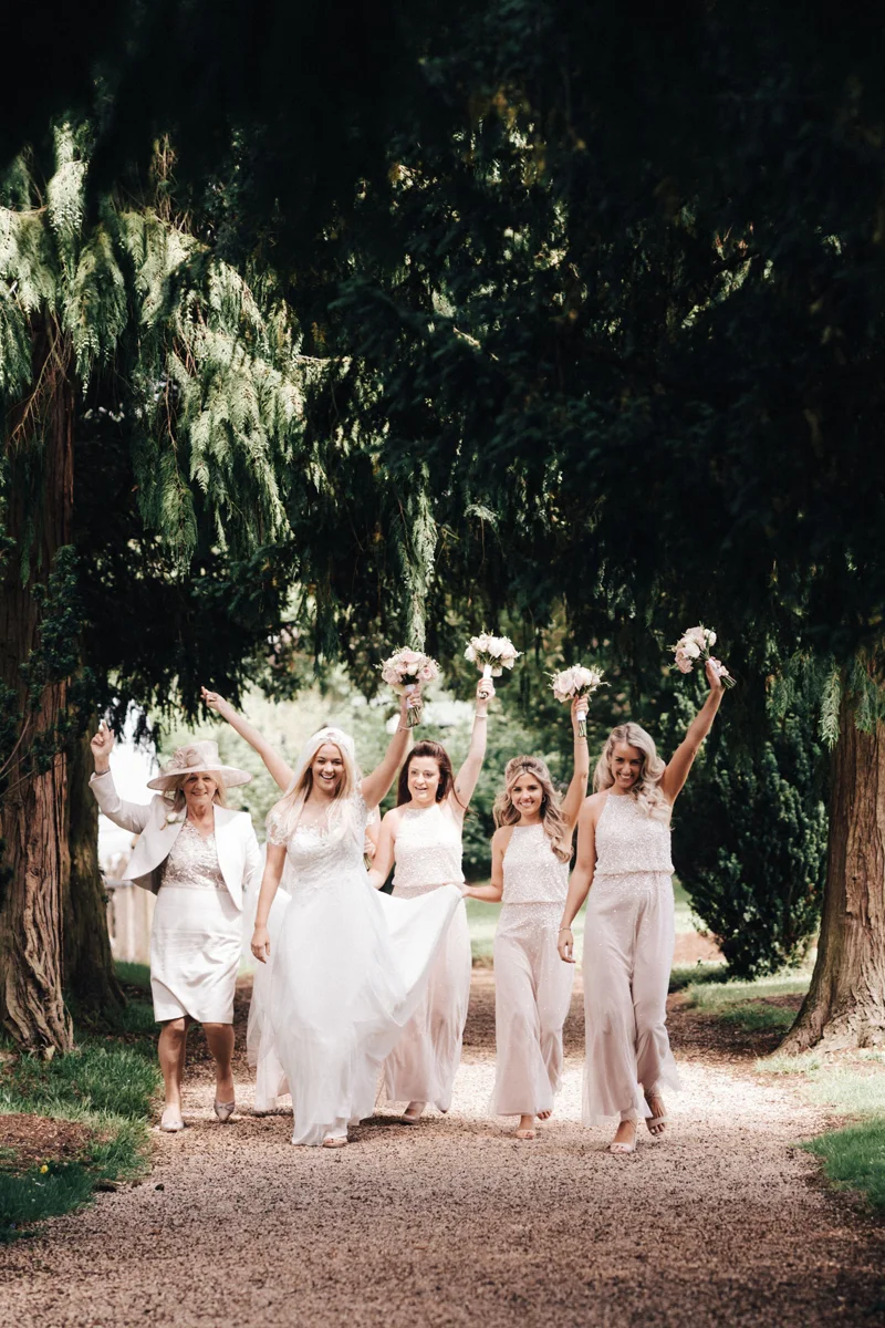 Bridesmaids and mother of bride walk up the path to the church holding bouquets in the air happily