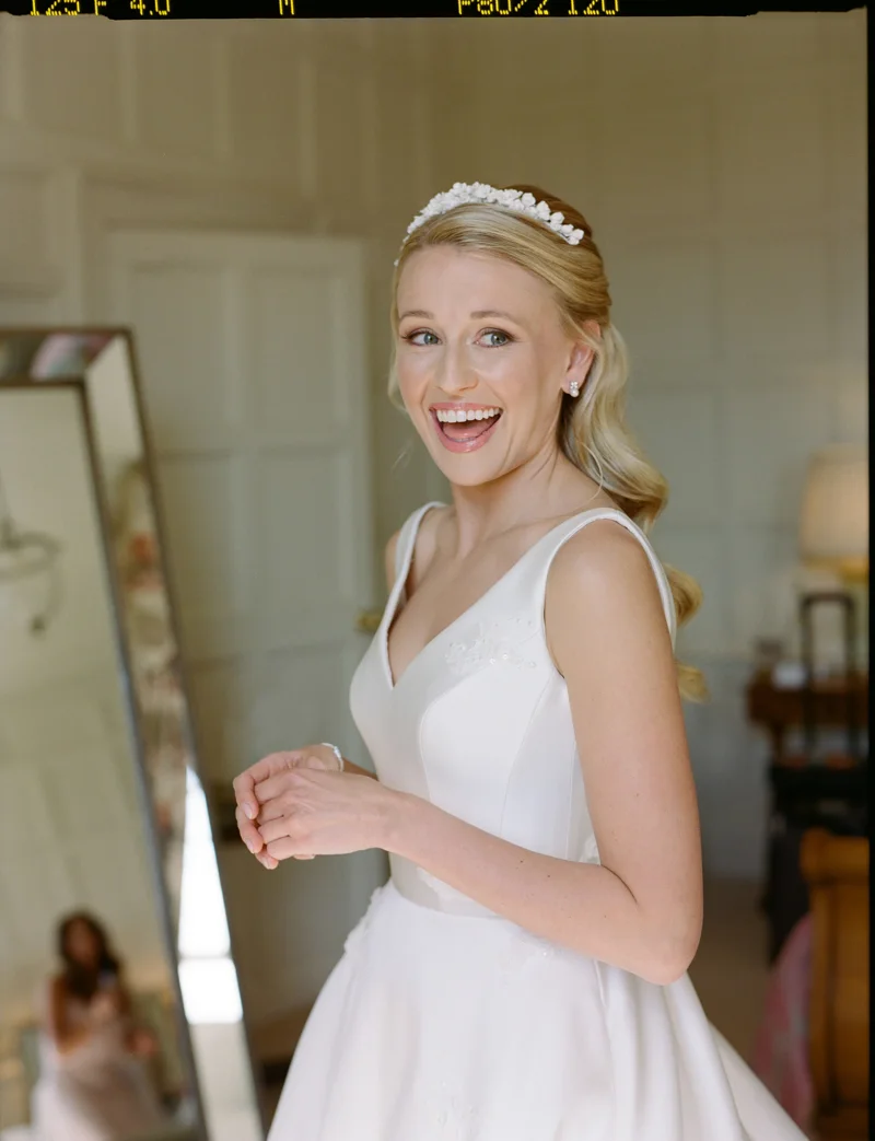 Elegant bride in simple satin dress with full skirt, pearl tiara and pearl earrings smiles on her wedding day at luxury Cotswolds wedding venue Elmore Court