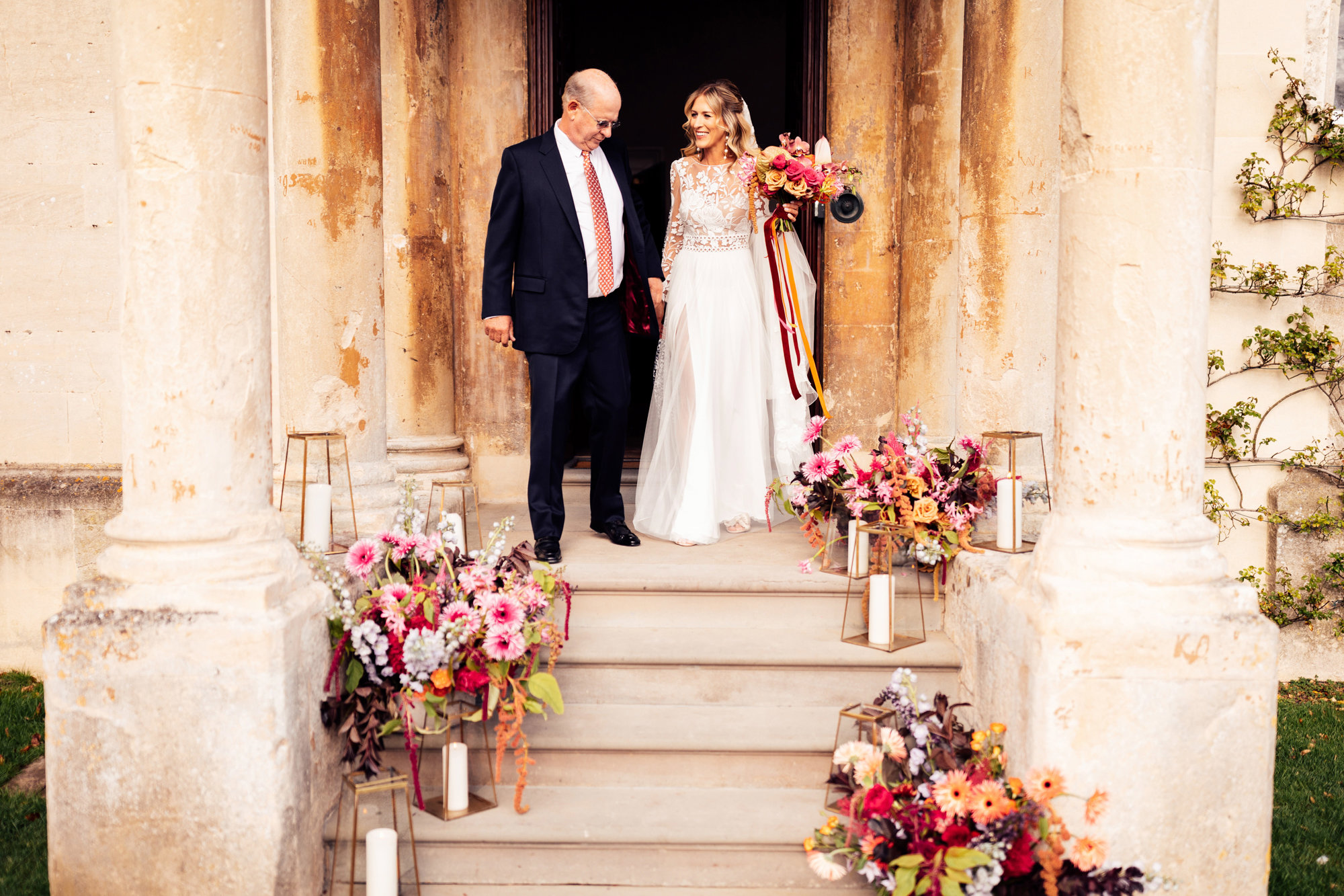 bride with father walking down step of mansion house decorated with colourful flowers for her wedding