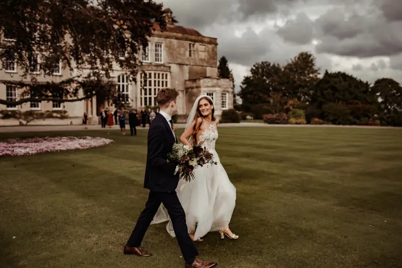 Modern boho bride and groom walk across the lawn in front of mansion house at their autumn wedding in October