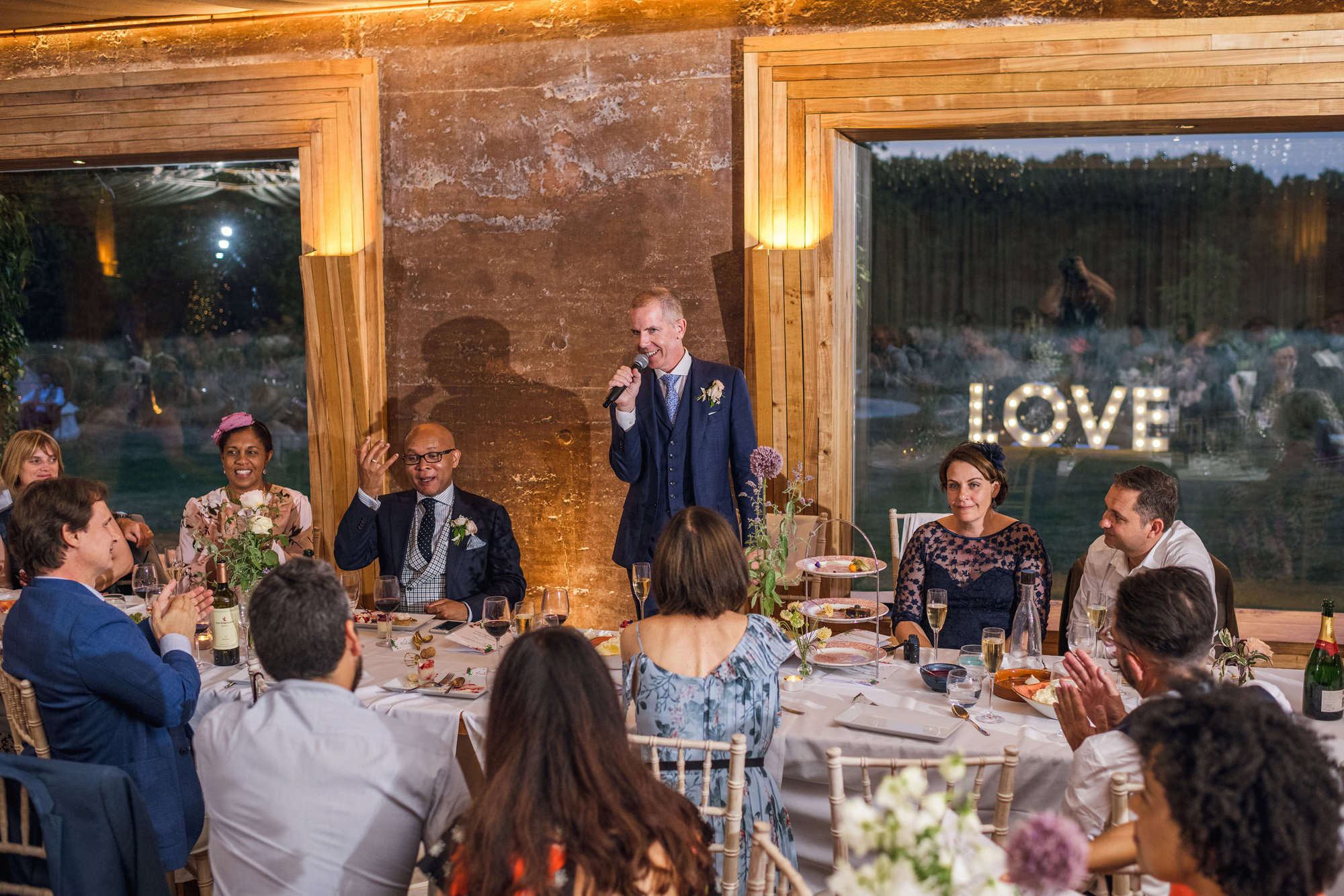 Gay wedding speeches, one groom makes speech whilst other listens inside rammed earth wedding venue with love light up letters seen through the window
