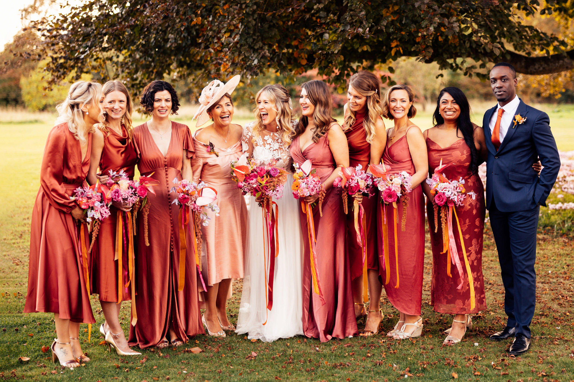 Colourful bridesmaids in orange dresses stand with boho bride with bright bouquets with ribbons in the cotswolds countryside