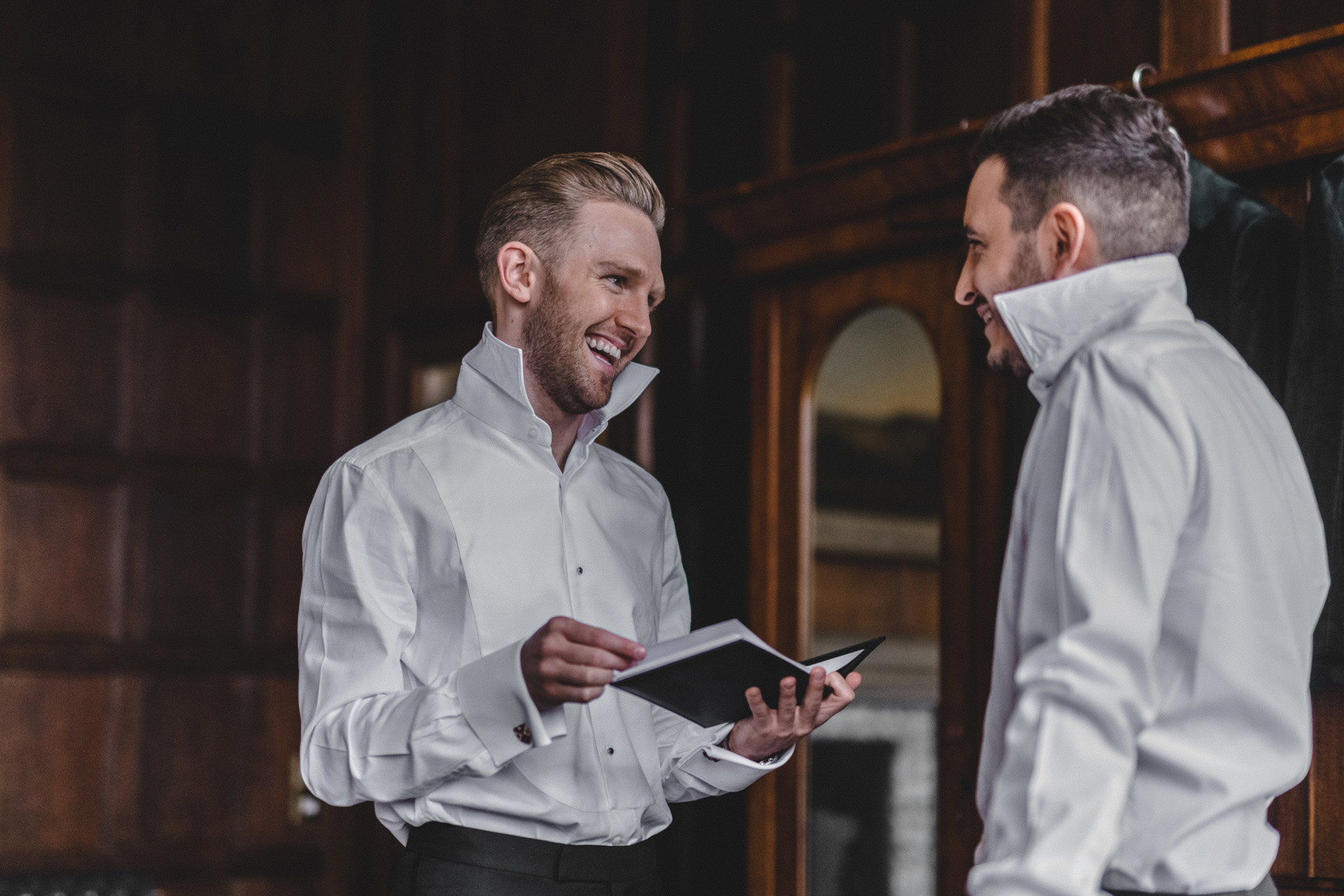 Two grooms in un buttoned shirts smile at each other as they get ready for their gay wedding