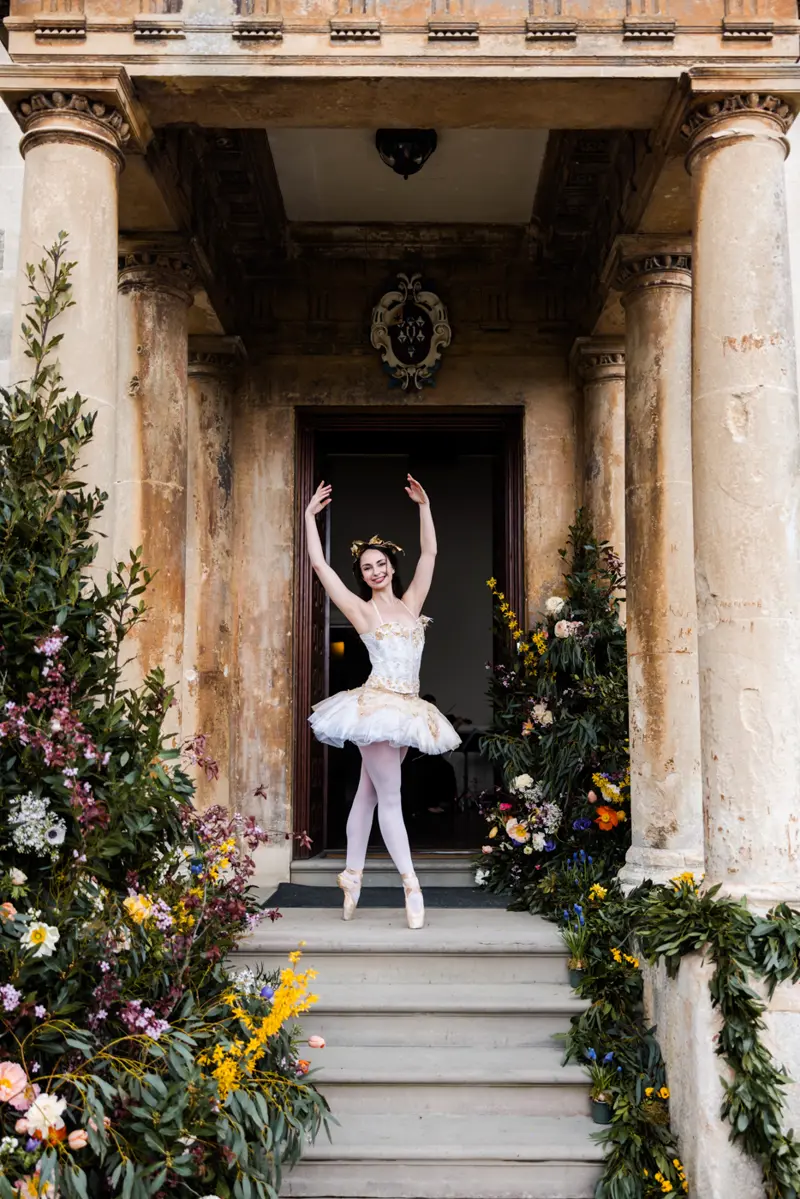 Divine company festival walkabout entertainment ballerina in white stands on steps with columns at stately home elmore court at unusual festival style wedding fair in gloucestershire