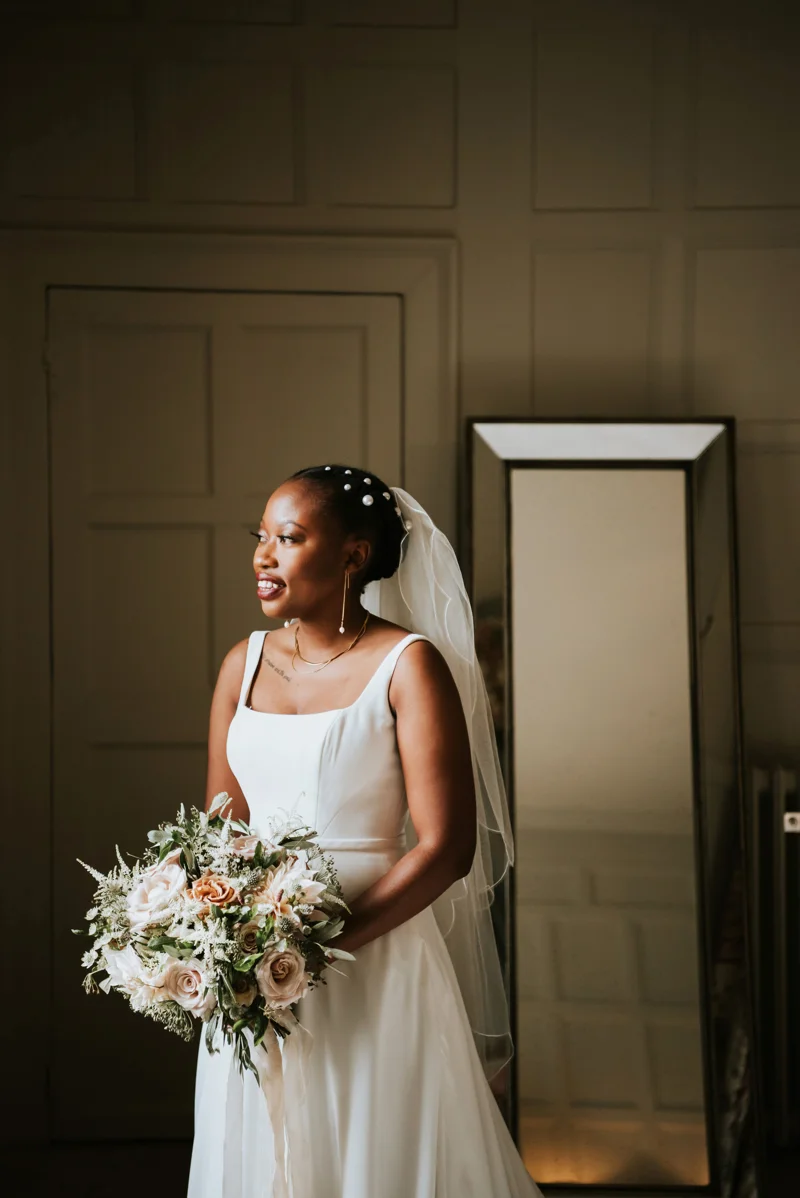Modern black bride wearing simple satin dress with pearl earrings and pearl hair pins holding chic bouquet of pink roses and greenery