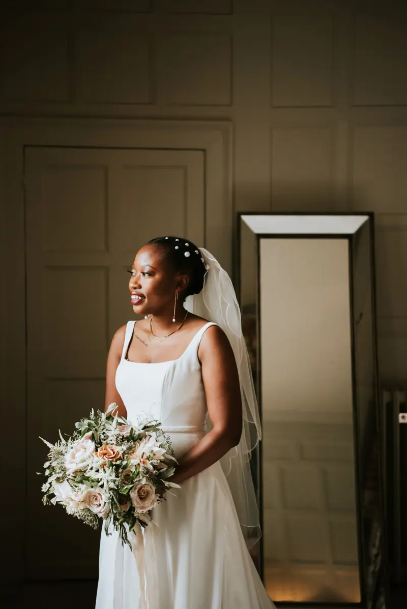 Modern black bride wearing simple satin dress with pearl earrings and pearl hair pins holding chic bouquet of pink roses and greenery