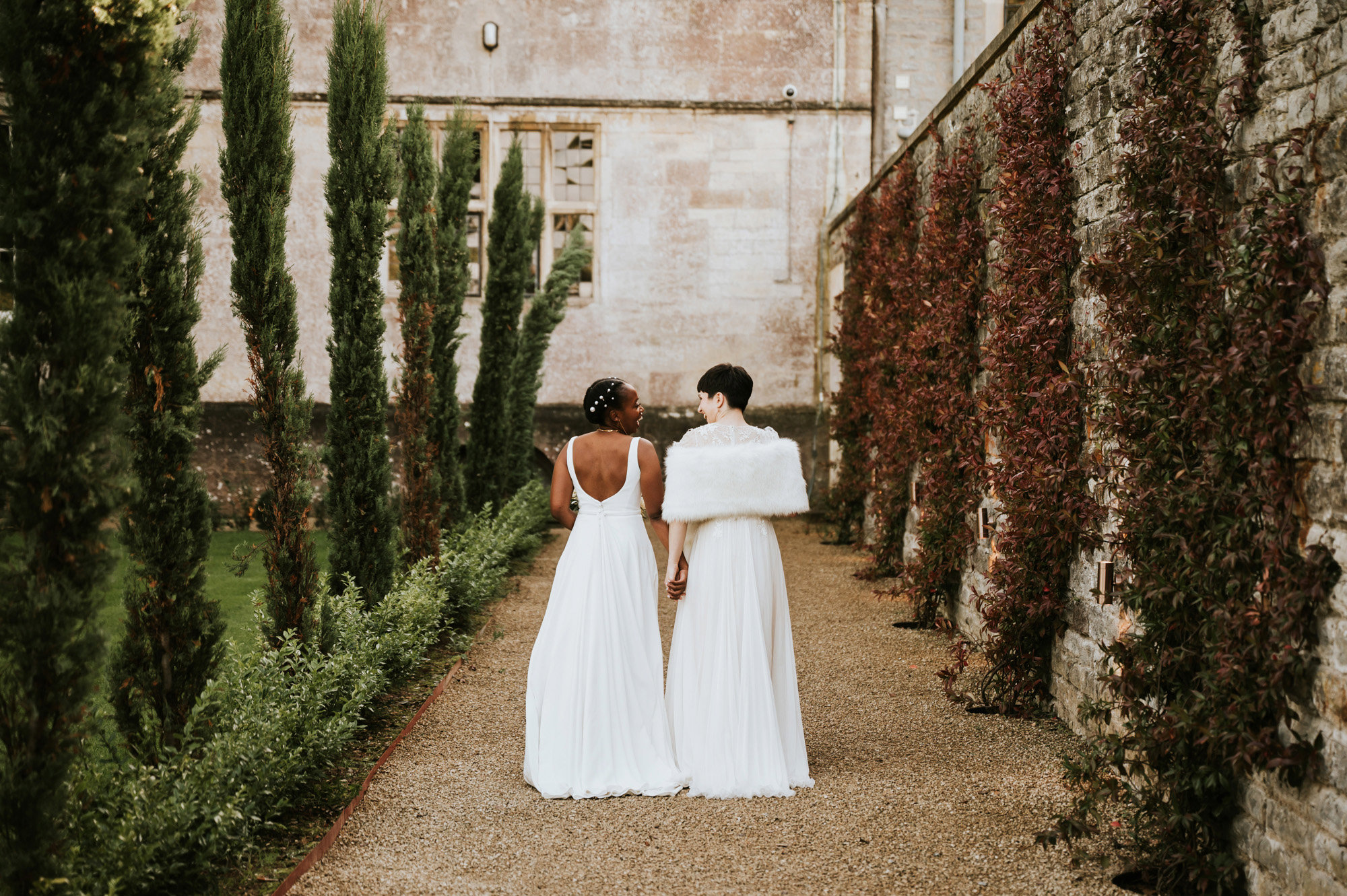 Lesbian wedding couple wearing floor length white wedding dresses with backs to camera walk serenely down tree lined path to their stately home wedding venue