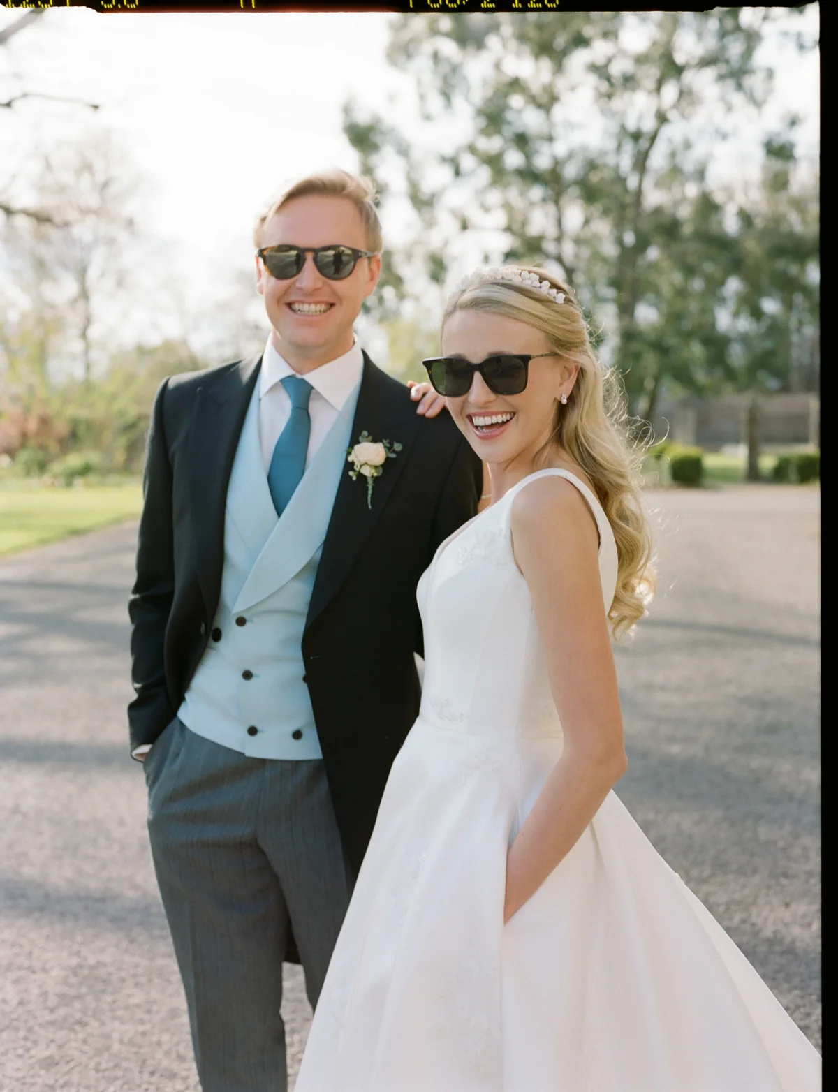 Cool bride wearing simple satin wedding dress with pockets, pearl tiara and pearl earrings and groom stand together in matching sunglasses