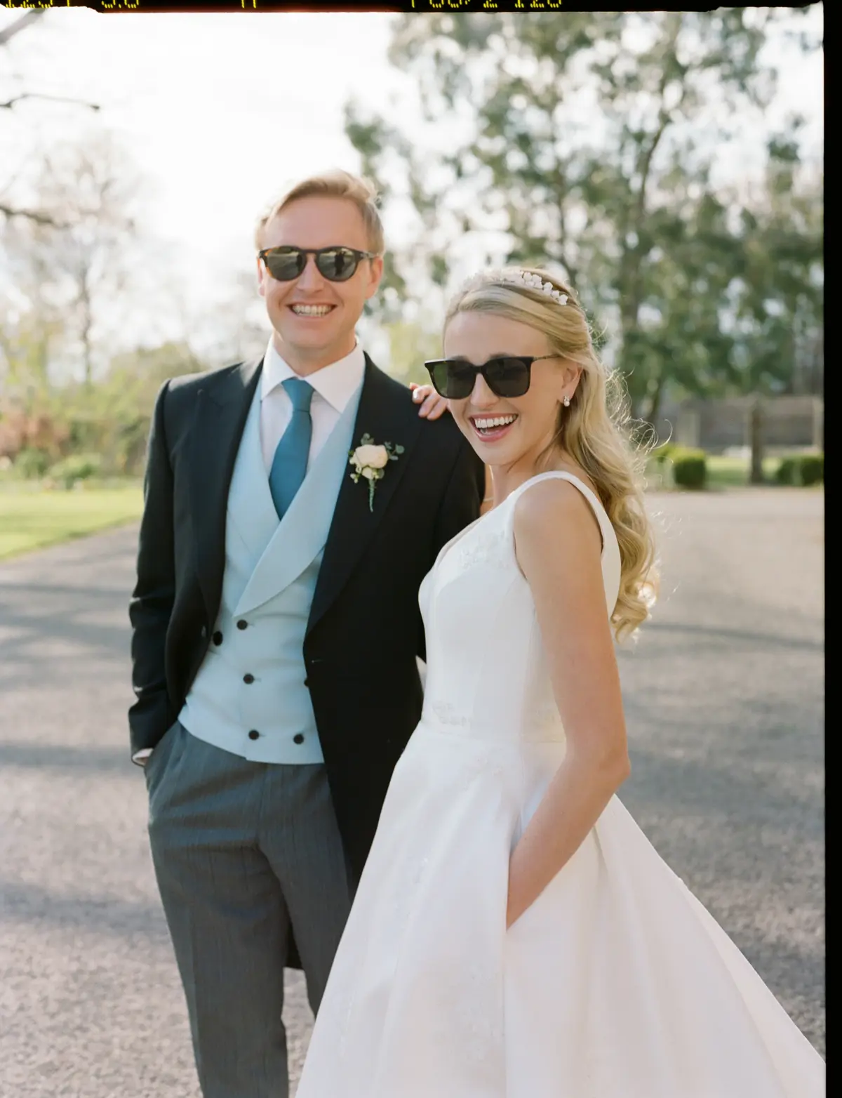 Cool bride wearing simple satin wedding dress with pockets, pearl tiara and pearl earrings and groom stand together in matching sunglasses
