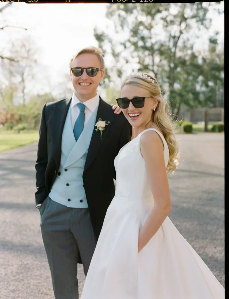 Cool bride wearing simple satin wedding dress with pockets, pearl tiara and pearl earrings and groom stand together in matching sunglasses